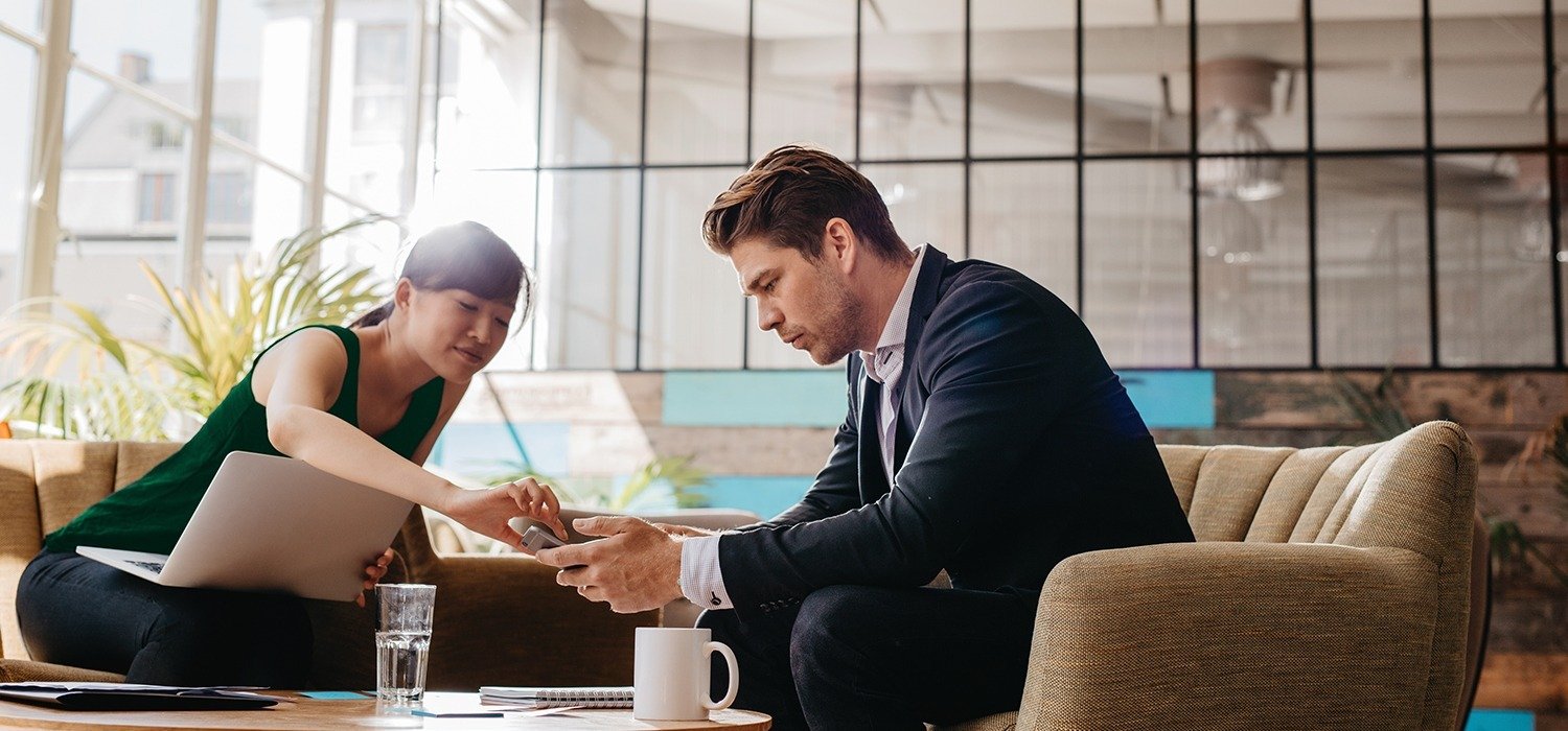 Professional and woman sitting together in loft office space. Woman is pointing at man's phone.