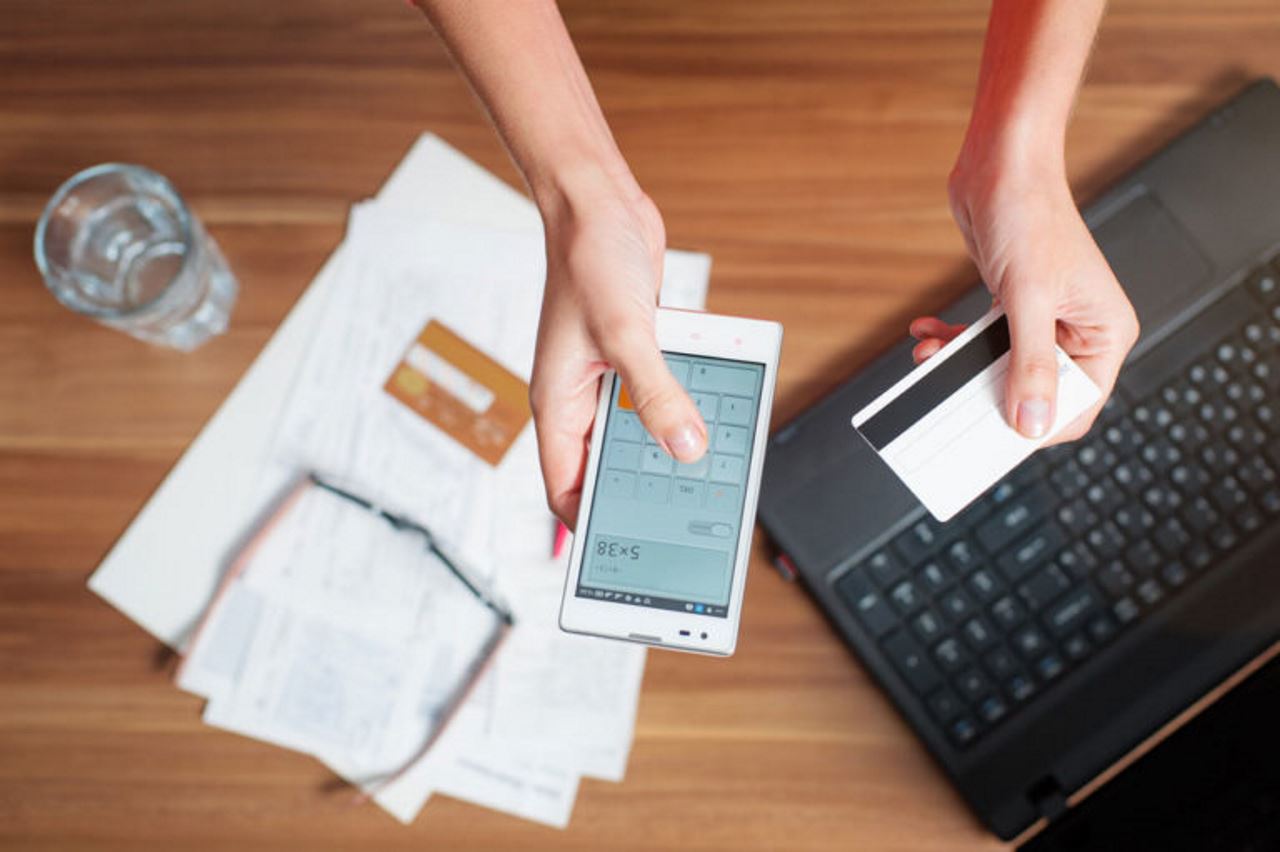 hands using calculator on phone holding credit card at desk over papers and laptop