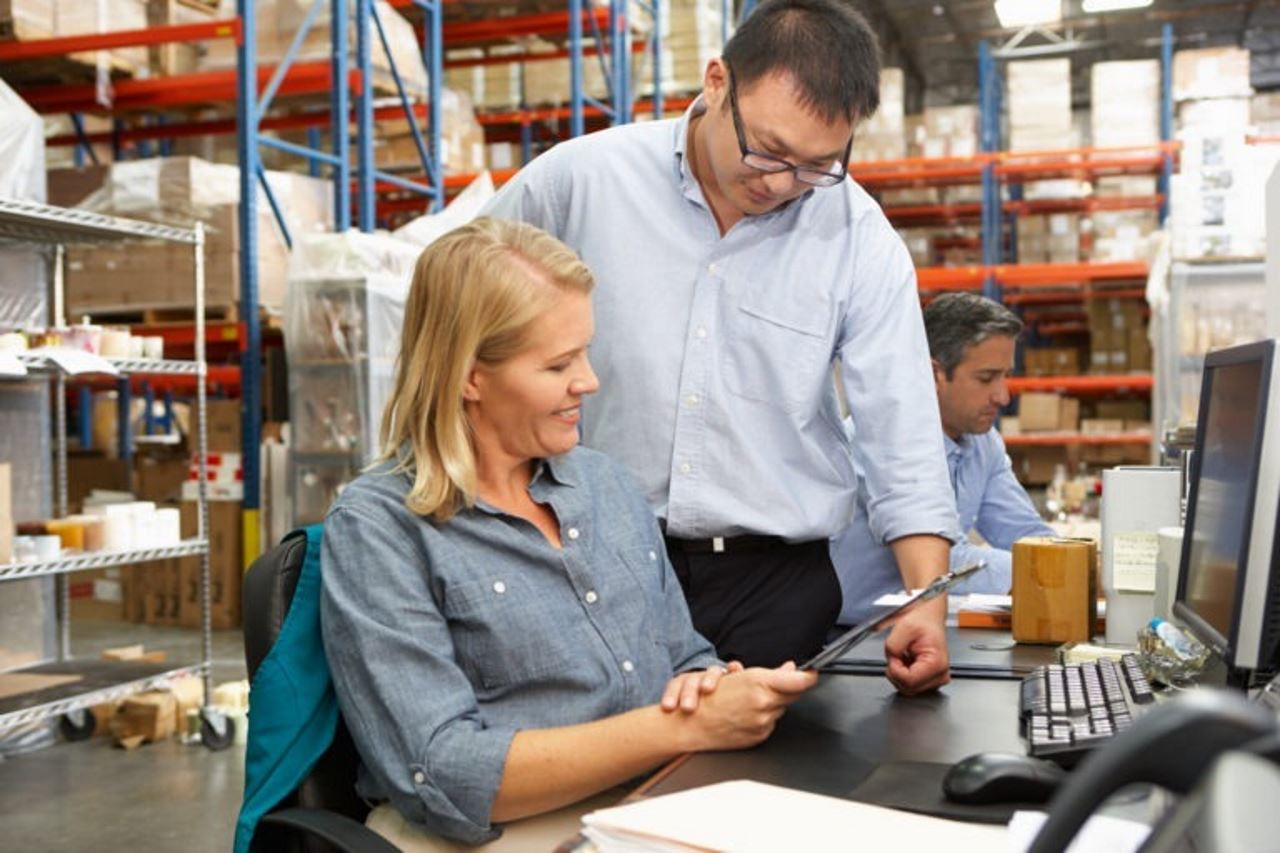Asian man and caucasian woman sitting at desk in warehouse looking at tablet together