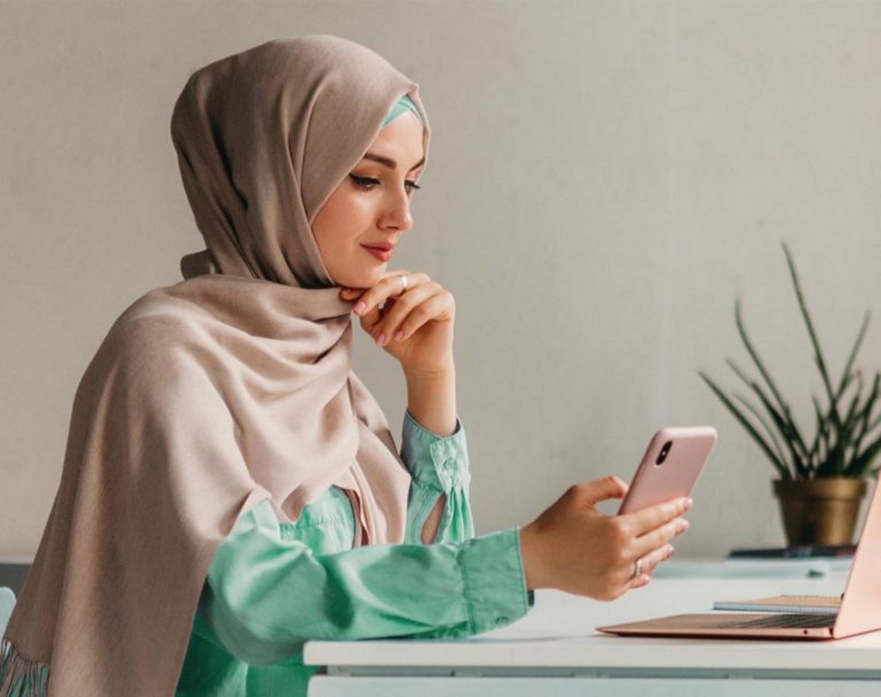 woman in home office sitting at table in front of laptop looking at cellphone