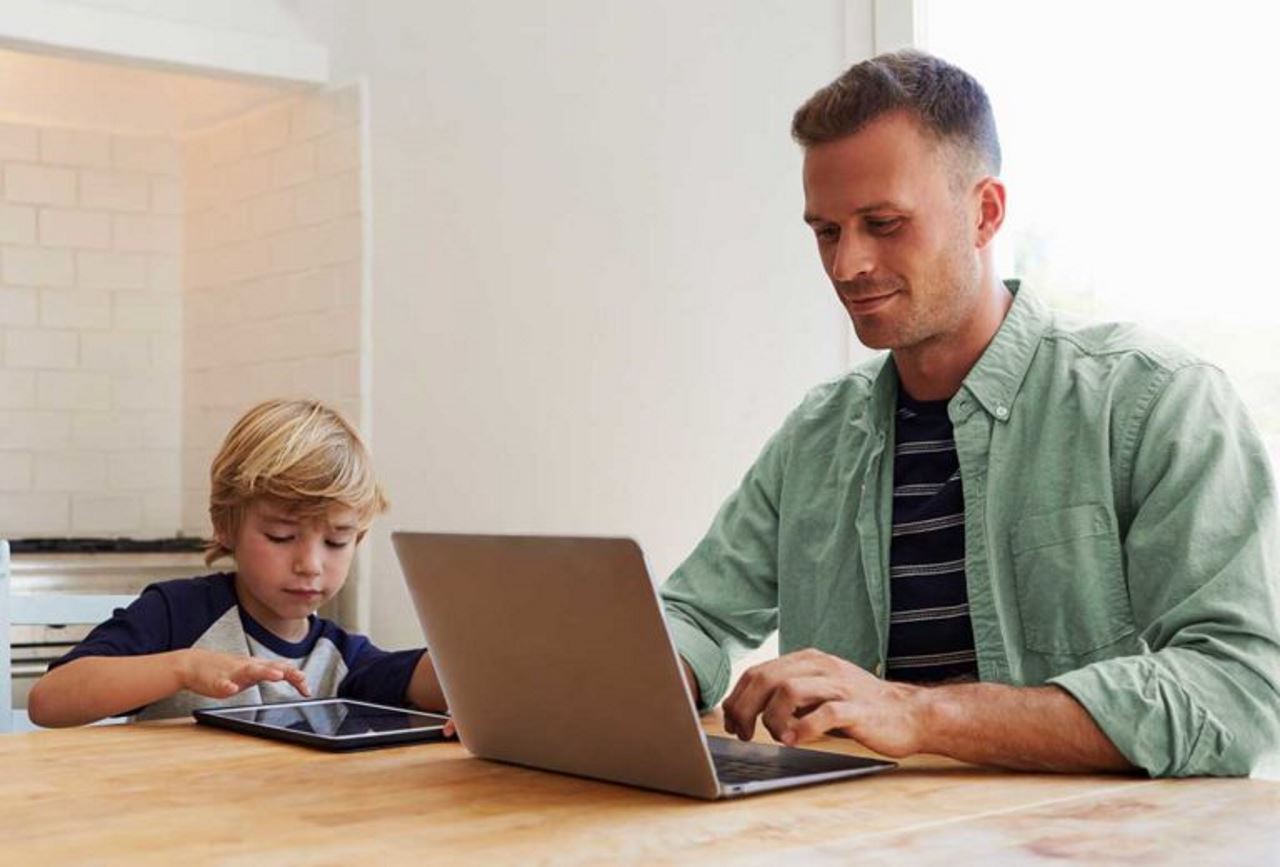 man in home office working on laptop while son plays on tablet beside him