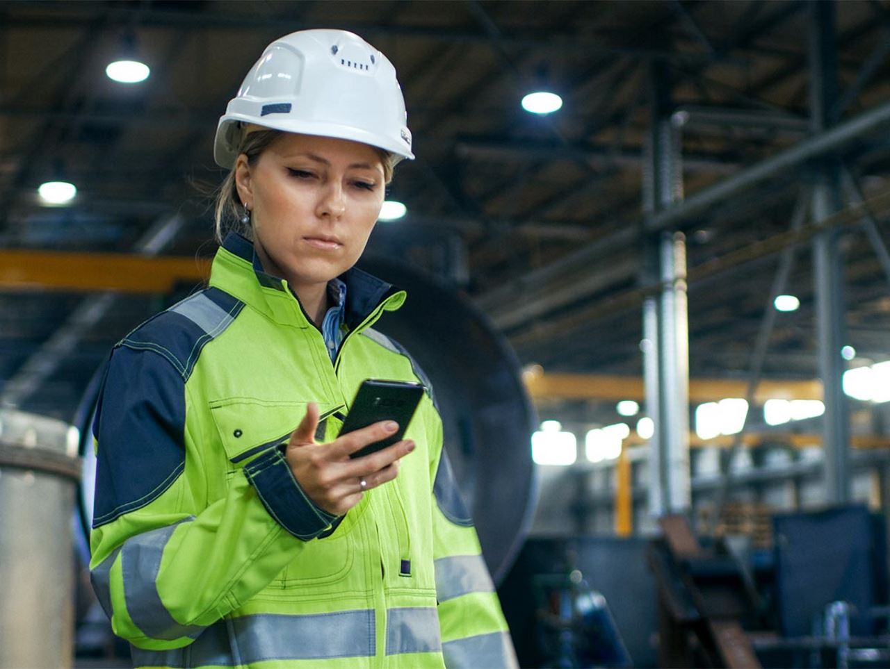 woman in warehouse with a hard hat looking at a cellphone