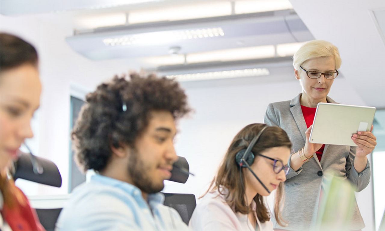 three employees sitting at work desk in Billtrust office with headsets while project manager watches over them with tablet