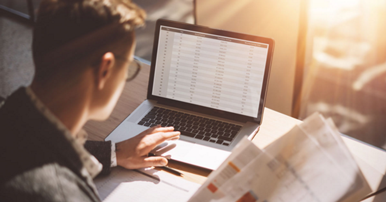 man sitting in front of laptop looking at a spreadsheet and holding stack of paper in one hand