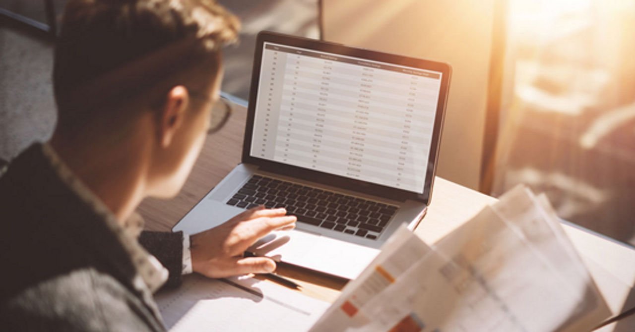  man sitting in front of laptop looking at a spreadsheet and holding stack of paper in one hand