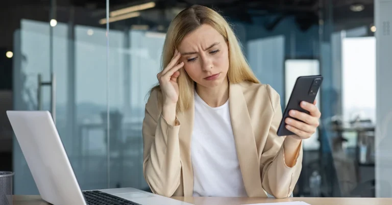 woman holding phone sitting at desk in front of laptop in office