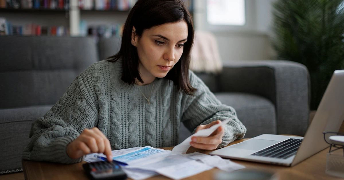 woman wearing sweatshirt holding invoice using calculator in front of laptop at desk