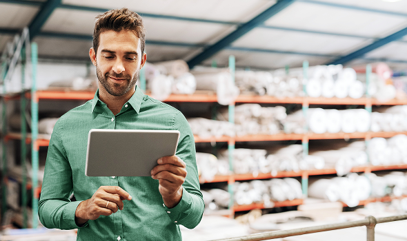 Eine Person hält ein Tablet vor eine Reihe von Regalen, die mit Rollen eines unbekannten Produkts gestapelt sind