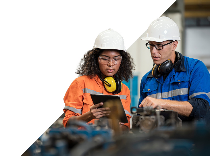 Een man en een vrouw in veiligheidsjumpsuits, beiden met een helm, een veiligheidsbril en gehoorbescherming, bekijken informatie op een tablet in een industriële omgeving.