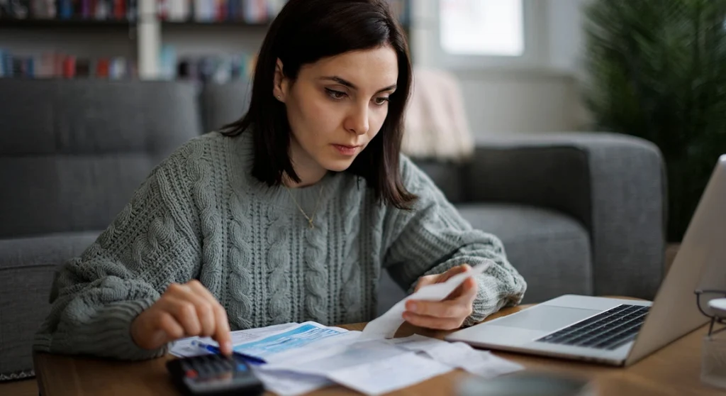 woman wearing sweatshirt holding invoice using calculator in front of laptop at desk