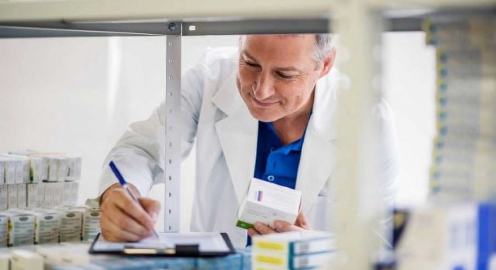 a pharmacist writing notes on a pad while performing inventory on a shelf filled with varied medications