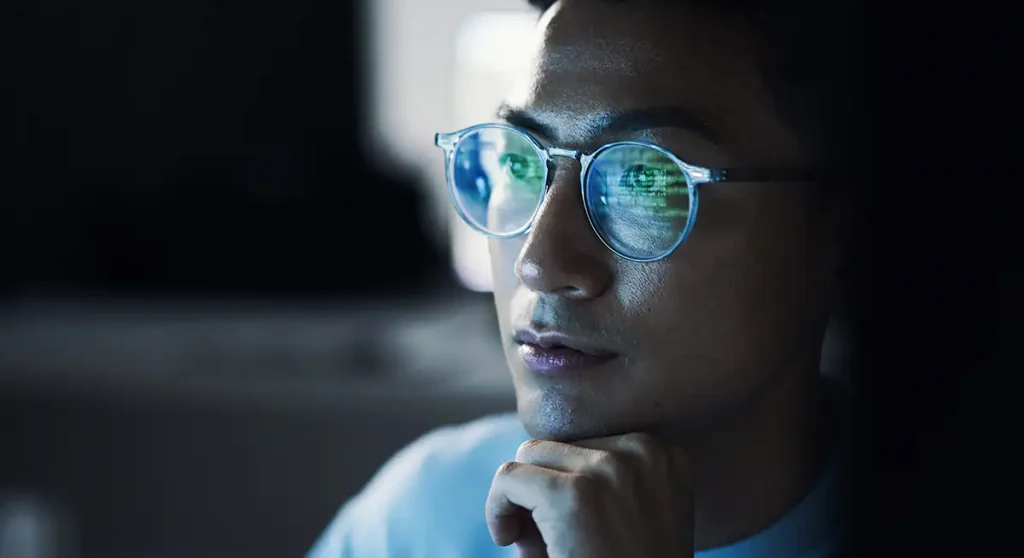 young man looking at computer in dark room with code reflected in glasses