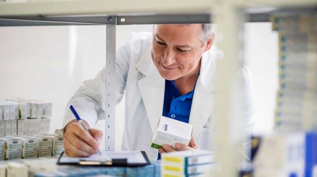 a pharmacist writing notes on a pad while performing inventory on a shelf filled with varied medications